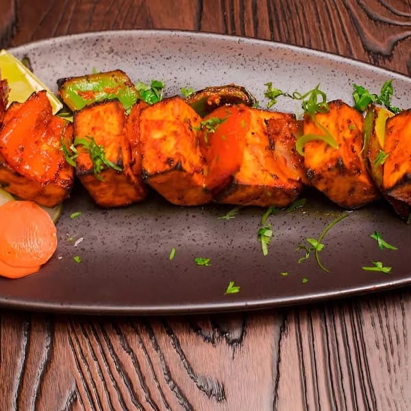 Grilled paneer tikka with green peppers, lime, pickled onions, and a side salad on a dark plate.