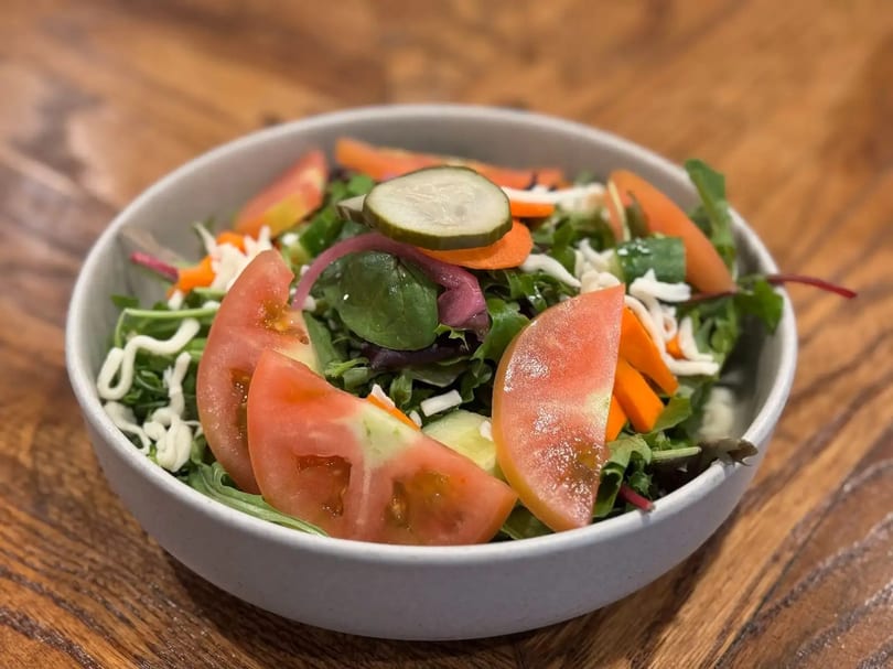 Close-up of a fresh garden salad with tomatoes, carrots, pickles, and dressing in a white bowl.