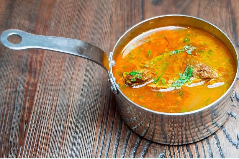 Hearty orange soup with meat and green herbs in a metal pot on a wooden table.