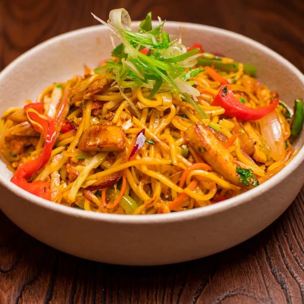 A bowl of vibrant stir-fried noodles with chicken, red peppers, and green onions on a wooden table.