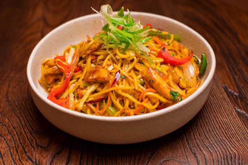 A bowl of vibrant stir-fried noodles with chicken, red peppers, and green onions on a wooden table.