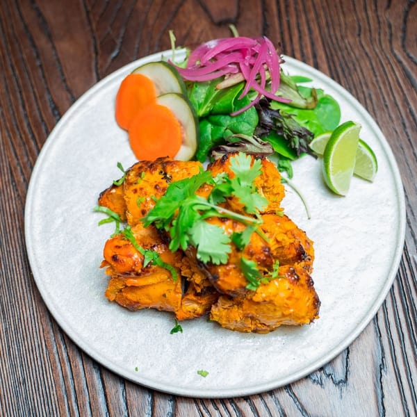 Tandoori chicken tikka with cilantro, pickled onions, salad, and lime wedges on a white plate.