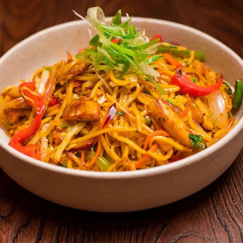 A bowl of chicken chow mein with noodles, vegetables, and red peppers on a wooden table.