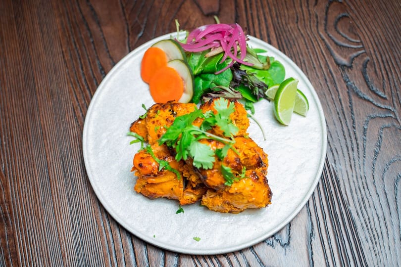 Tandoori chicken tikka with cilantro, pickled onions, carrots, cucumbers, and lime wedges on a white plate.